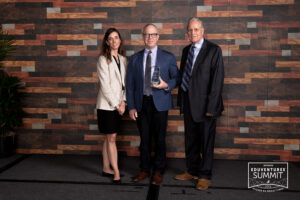 A woman and two men stand in front of a brick wall. The man in the middle is holding a plaque and smiling.